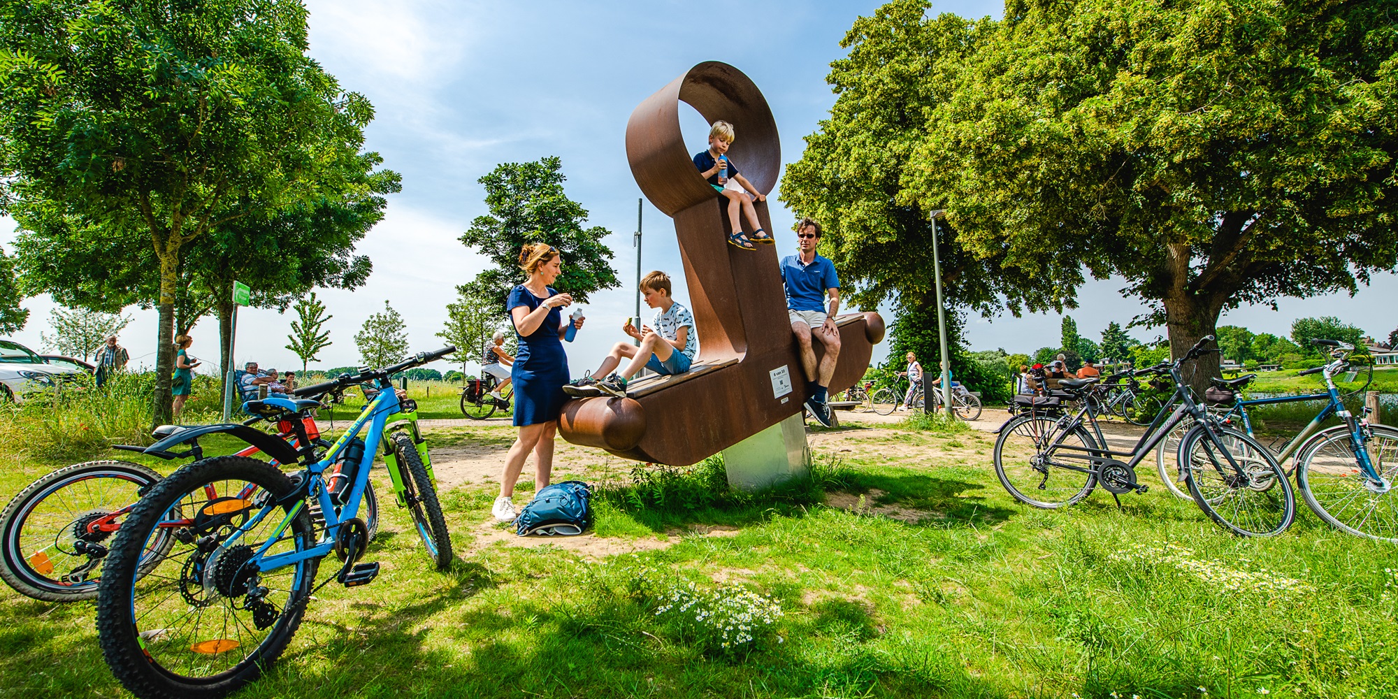 Fietsers en een gezin nemen pauze bij het kunstwerk ‘drakenzwaard’ in Beesel langs de Maasfietsroute, zittend op het grote metalen zitobject te midden van een groen, zomers landschap.