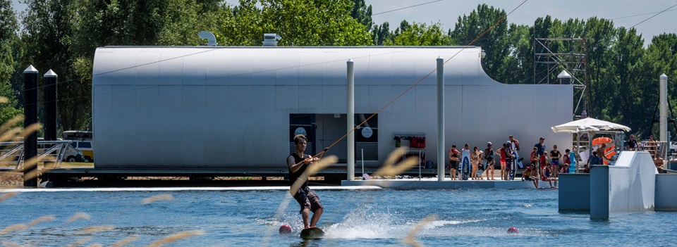 Wakeboarder in actie op het water bij Beaver Creek, met toeschouwers en een modern gebouw op de achtergrond op een zonnige dag.