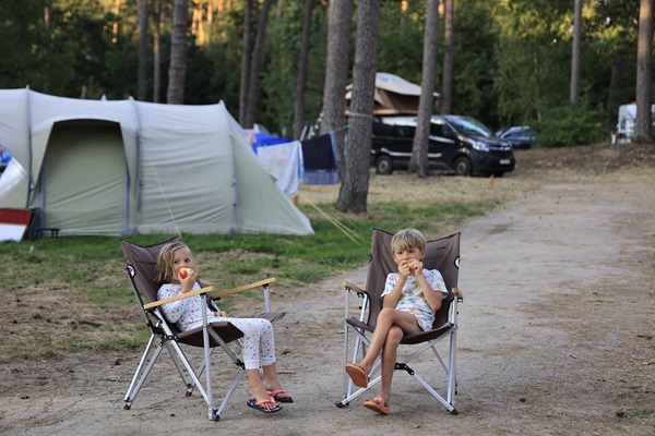 Zwei Kinder sitzen in Campingstühlen und essen einen Apfel auf einem Campingplatz bei Huttopia in der Gemeinde Roerdalen, mit Zelten und Bäumen im Hintergrund.